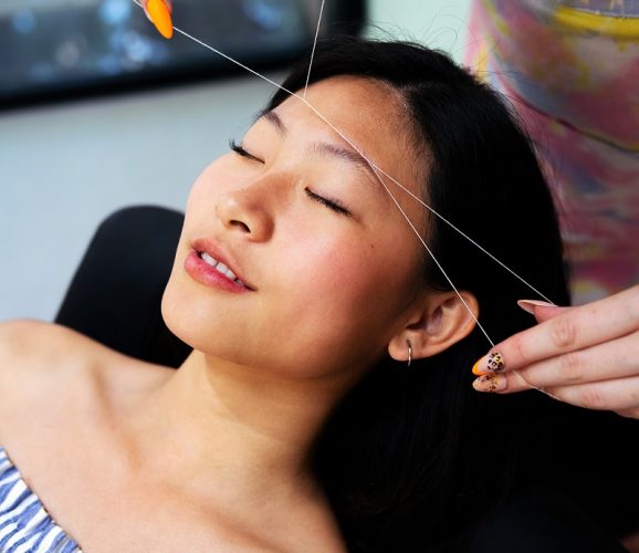 professional beautician performs an eyebrow threading on a young asian woman in the beauty salon, concept of wellness and body care