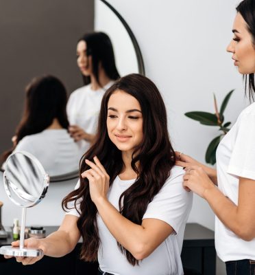 Beautiful woman hairdresser is making hairdress on a adorable woman client in a hair styling salon indoors. Pleasure lady looking at the hand mirror