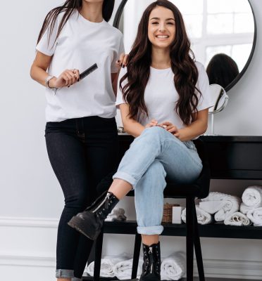 Full length view of the hairdresser making hairstyle for young woman in beauty salon. Two cheerful girls looking at the camera with pleasure smiles